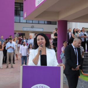 Okul bahçesinde mor kürsüde mikrofonla konuşan beyaz ceketli kadın, yanında takım elbiseli bir erkek