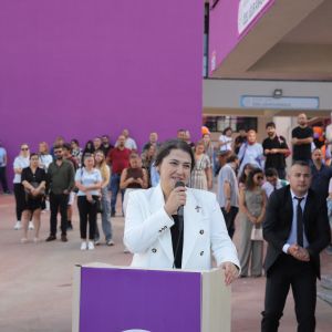 Mor kürsünün başında konuşan beyaz ceketli kadın, yanında takım elbiseli bir erkek, arkada izleyen kalabalık