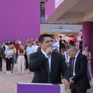 Okul bahçesinde mor kürsüde mikrofonla konuşan gözlüklü, takım elbiseli erkek, yanında bir erkek, arkada dinleyen kalabalık