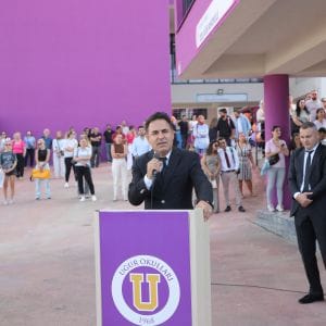 Uğur Okulları armalı mor kürsüde mikrofonla konuşan takım elbiseli erkek, yanında ayakta bekleyen bir erkek